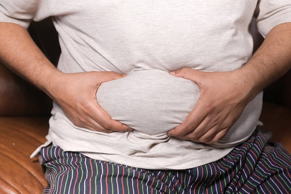 A close-up view of a man holding his tummy while sitting on a couch, highlighting lifestyle choices.