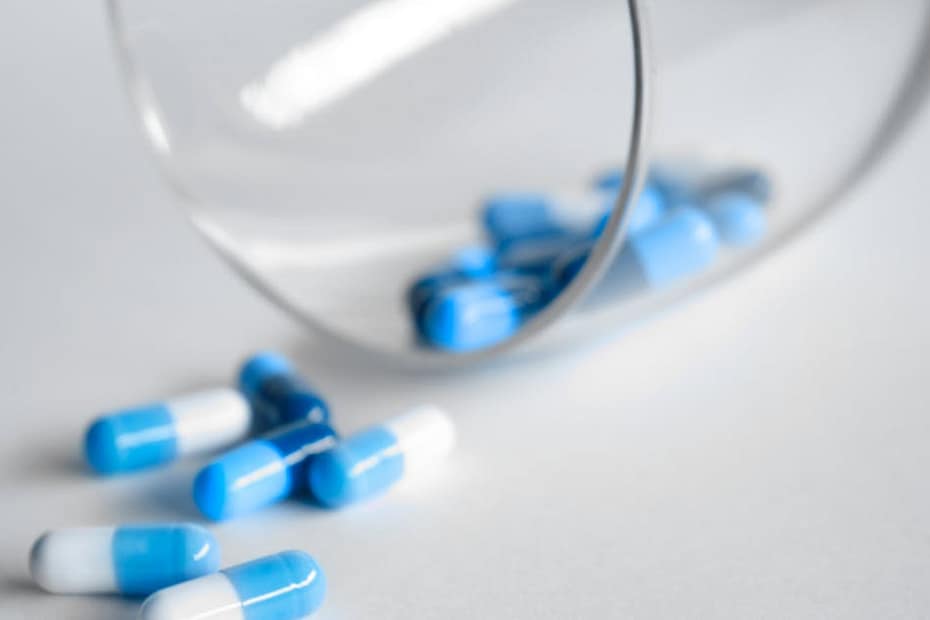 A close-up of blue and white capsules spilling from a glass on a light background.