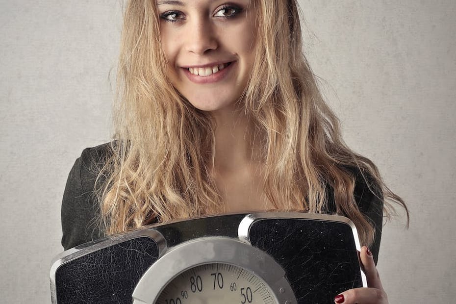 Young woman with blond hair smiling, holding a vintage bathroom scale indoors.