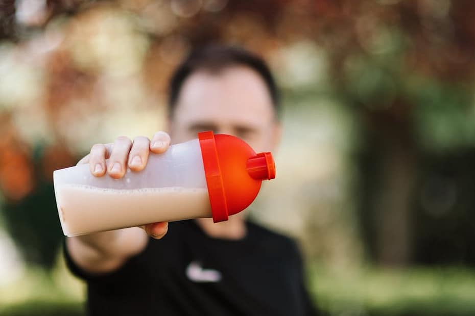 Focus on a man holding a plastic protein shake tumbler outdoors with a blurred background.