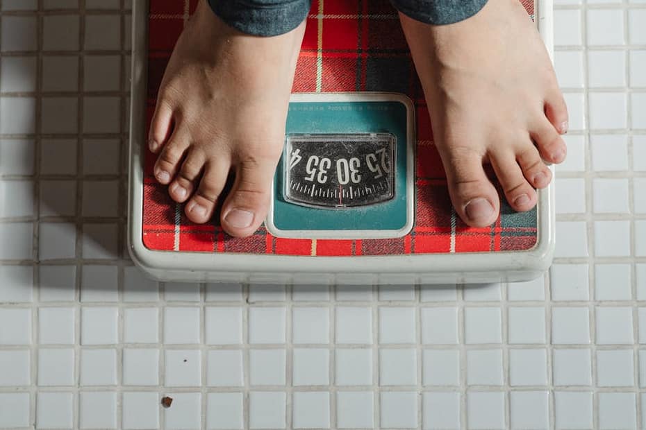From above crop anonymous barefoot child in jeans standing on weigh scales on tiled floor of bathroom