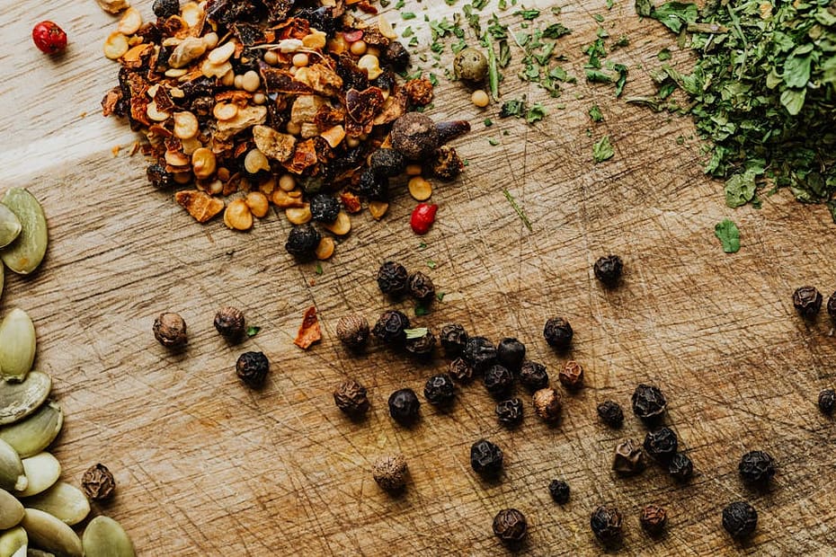 A close-up of various herbs and spices including peppercorns and pumpkin seeds on a rustic wooden surface.