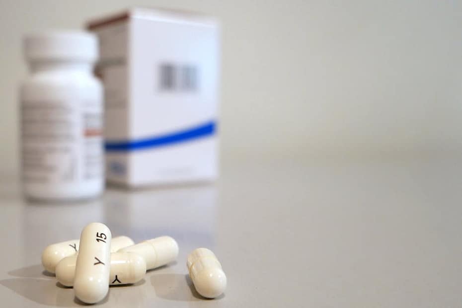Close-up image of white capsules with medicine bottle and box in the background.