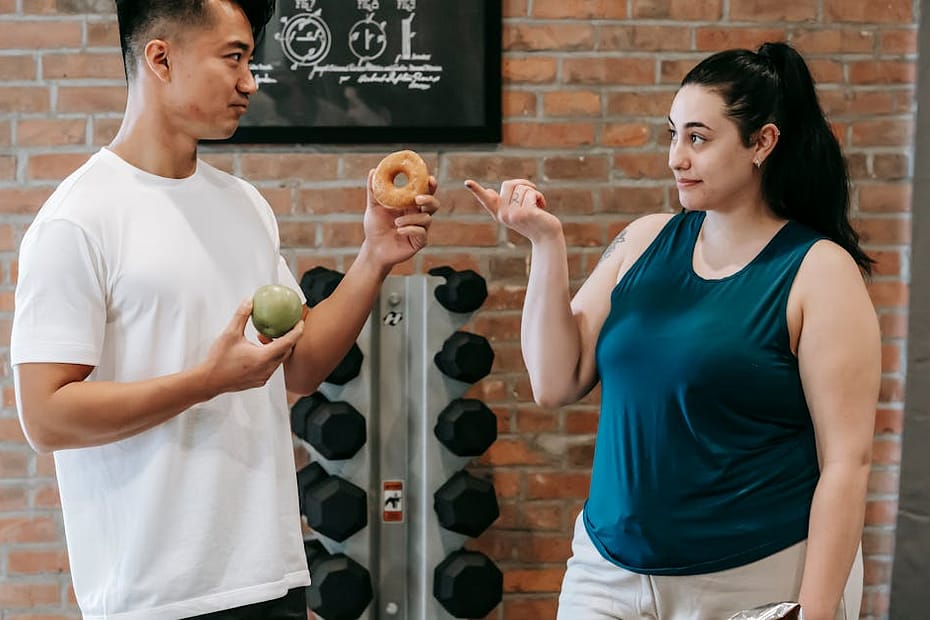 Side view of Asian personal instructor with apple and doughnut offering healthy food to plus size woman while standing in gym