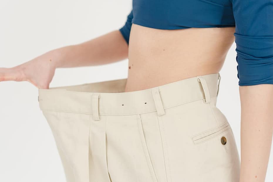 A woman demonstrates significant weight loss by wearing oversized pants indoors.