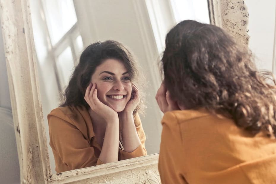 A cheerful woman smiles at her reflection in a vintage-style mirror, exuding positivity and warmth.