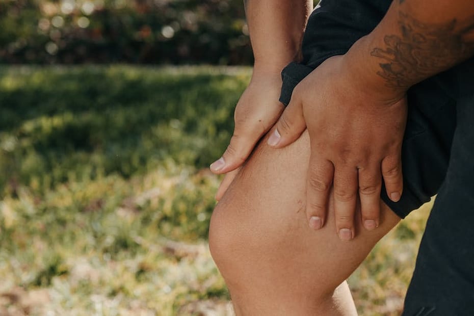 A close-up image of a person holding their knee, indicating pain or injury, with greenery in the background.