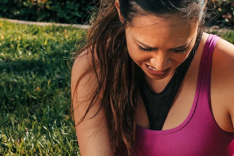 A young woman in activewear sitting on grass, massaging her sore knee in a park setting.