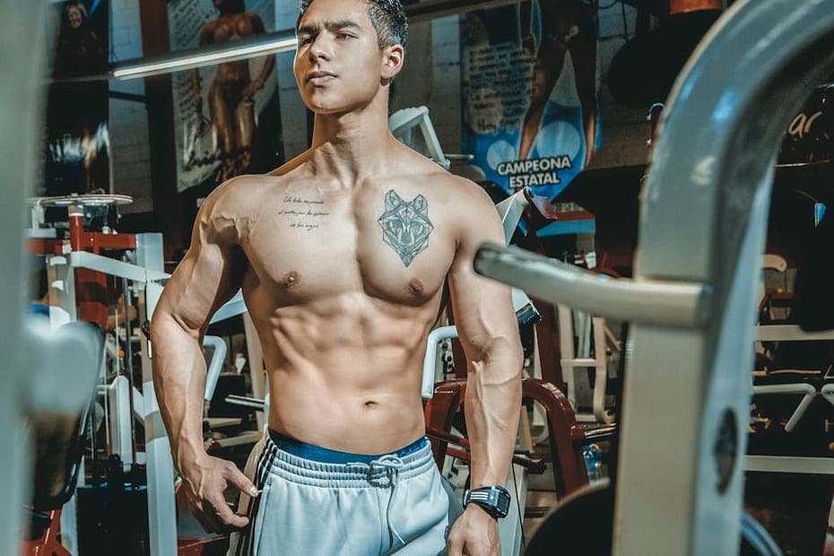 Shirtless man with tattoos posing in a gym environment, showcasing fitness and strength.