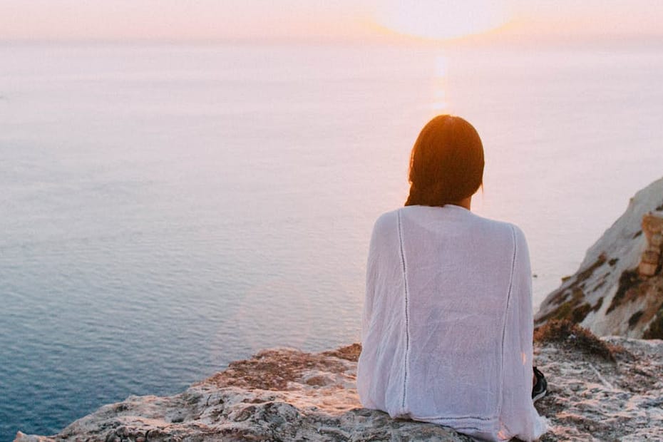 A serene view of a woman sitting on a rocky cliff, gazing at the ocean during sunset. Perfect for relaxation and travel themes.