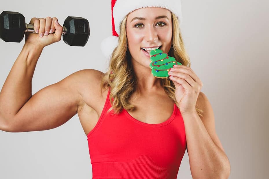 Woman in Santa hat lifting dumbbell and enjoying a holiday cookie, embracing festive fitness.