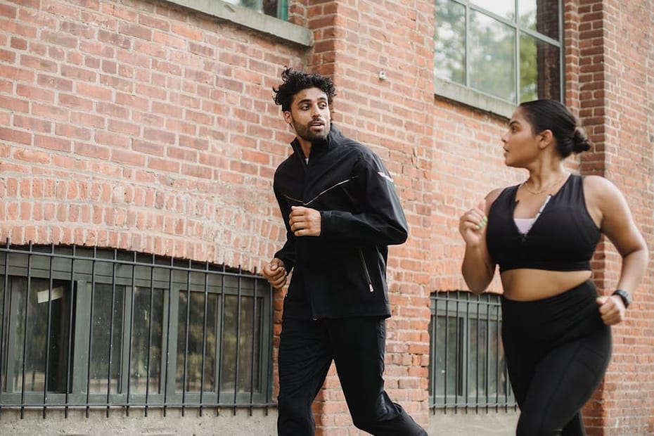 A man and woman in athletic gear jogging beside a brick wall outdoors, expressing energy and communication.