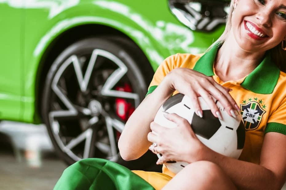 Smiling woman in sports attire hugging a soccer ball, sitting next to a vibrant green car indoors.